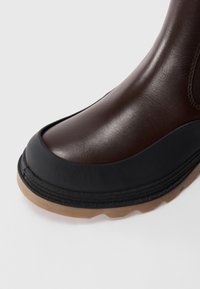 Botte en cuir marron avec un bout arrondi, dotée d'une protection en caoutchouc noir et d'une semelle noire texturée. Coutures visibles et finition douce mate.