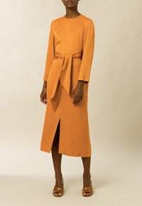 Vestido naranja de manga larga, hecho de una tela suave, con la cintura atada y una abertura frontal. Se llevan sandalias de tacón marrones y destalonadas.