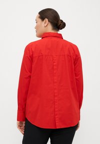 Camicia rossa con colletto, maniche lunghe e orlo arrotondato. Presenta un dettaglio a yoke sul retro e bottoni nascosti lungo la schiena.