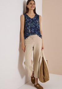Femme debout contre un mur, portant un haut sans manches bleu à fleurs, un pantalon beige à jambes larges, des chaussures marron, et tenant un grand sac fourre-tout tressé.