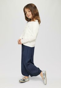 Biała, długosleeve kardigan z falbaniastym dołem, zestawiony z granatowymi, plisowanymi spodniami oraz błyszczącymi, srebrnymi balerinami z kokardkami.