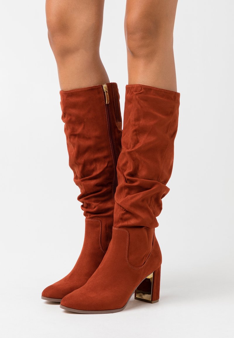 Botas de gamuza de color óxido, hasta la rodilla, con un diseño desgarbado, punta afilada y un tacón cuadrado con acentos dorados, que cuentan con un cierre lateral.