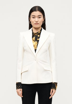 Frau trägt weißen maßgeschneiderten Blazer über schwarzem und gold gemustertem Hemd, kombiniert mit schwarzen Hosen, vor einfarbig hellem Hintergrund stehend.