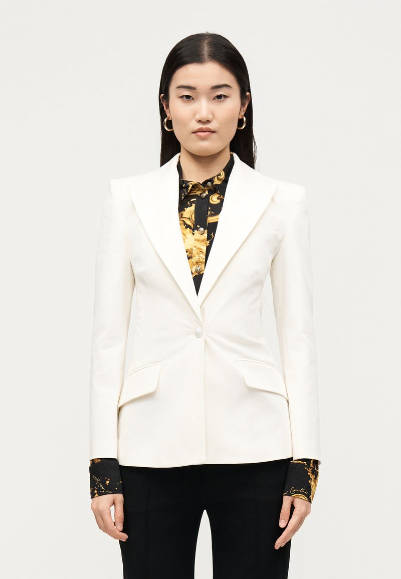 Γυναίκα φοράει λευκό προσαρμοσμένο σακάκι πάνω από μαύρο και χρυσό πουκάμισο με σχέδια, συνδυασμένο με μαύρο παντελόνι, στέκεται μπροστά σε απλό ανοιχτό φόντο.