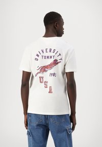 Vīrietis, kura mugurā ir balts T-krekls ar lecošu tīģeri un uzrakstu "University Tommy 1985 USA", apvienots ar zilām džinsu biksēm.