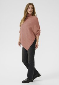 Poncho en tricot rose surdimensionné avec un col haut, doté d'un motif texturé, associé à un jean noir à jambes droites et des chaussures noires.