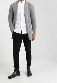 Cardigan grigio a coste sopra una camicia bianca con bottoni, abbinato a pantaloni neri e stivaletti neri. Tessuto liscio e colori neutri.