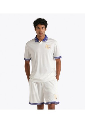 Giovane uomo che indossa una polo bianca e pantaloncini con rifiniture blu e un logo giallo di cartone animato, in piedi con le mani nelle tasche.