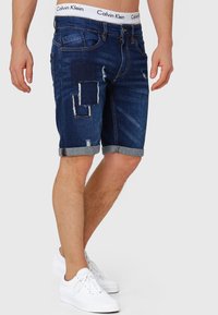 Tamno plavi traper šortsi s istrošenim mrljama, zavrnutim rubovima i pojasom s Calvin Klein logotipom. Udoban kroj s ravnim stajlingom.
