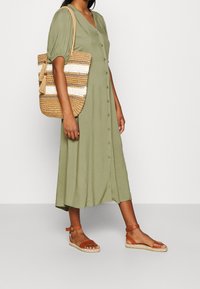 Olivgrünes Midi-Kleid mit Knöpfen, kombiniert mit braunen Riemchensandalen und einer geflochtenen Handtasche mit beigefarbenen und weißen Streifen.