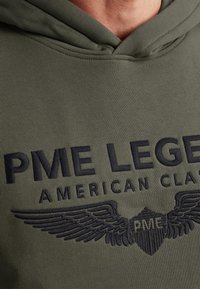 Zielona oliwkowa bluza z kapturem, z czarnym haftowanym napisem "PME LEGEND AMERICAN CLASSIC" oraz skrzydlatym wzorem na obszarze klatki piersiowej.