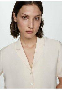 Blusa crema chiaro con maniche corte, frontale con bottoni e colletto a scollo a "V". Texture liscia e leggero lucido. I bottoni sono di colore chiaro.