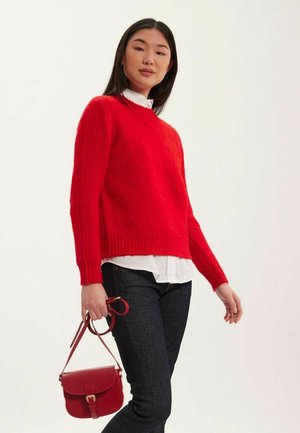 Un pull rouge en tricot avec un col rond et des poignets côtelés, associé à une chemise blanche, un jean sombre et un petit sac bandoulière rouge.