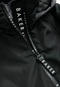 Veste noire avec une fermeture éclair dotée d'une tirette de marque. L'intérieur présente une texture matelassée, mettant en avant une surface extérieure lisse.