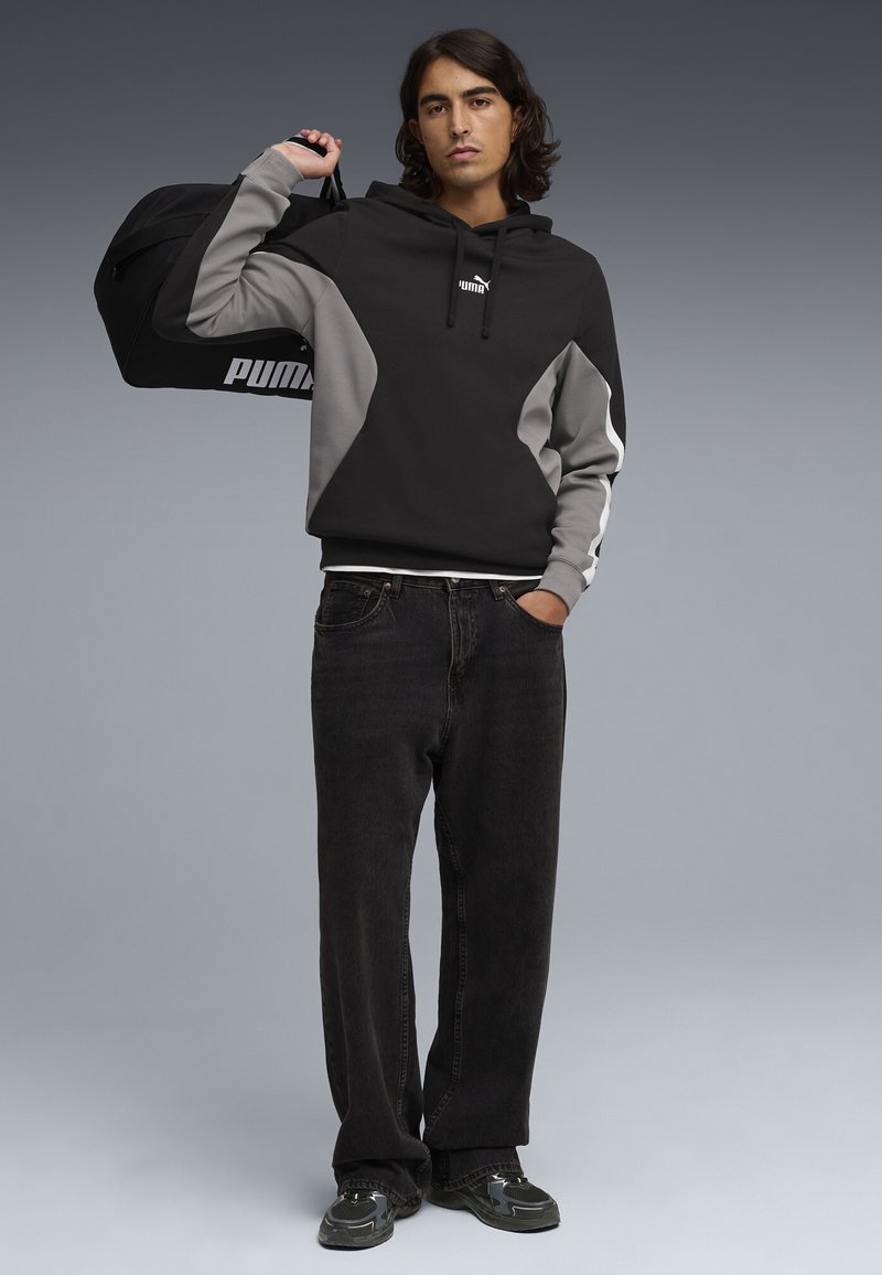 Jeune homme portant un sweat à capuche Puma noir et gris et un jean noir, tenant un sac de sport Puma noir sur l'épaule, sur fond gris.