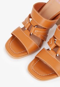 Brune læder åbne sandaler med hvid syning, to brede remme og en blokhæl på en hvid baggrund.
