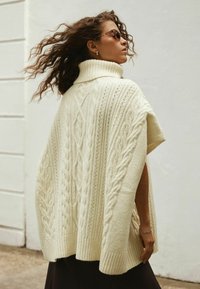 Cremfarbener, kabelgestrickter Poncho mit hohem Kragen und kurzen Ärmeln, der texturierte Muster und einen gerippten Saum aufweist. Trägt Sonnenbrillen.