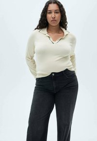 Light cream sweater with a collar and black trim, paired with high-waisted black denim jeans. Curly hair, neutral background.