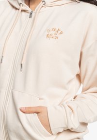 Felpa rosa chiaro con zip realizzata in tessuto morbido, caratterizzata da una tasca frontale, cappuccio con coulisse e logo ricamato con dettagli floreali.