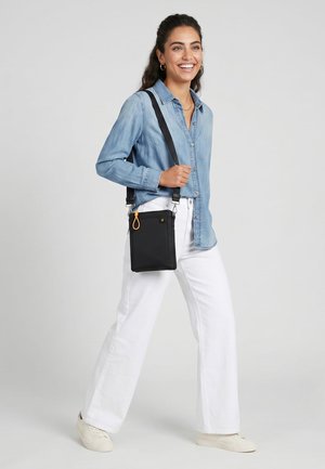 Kobieta idąca z czarną torebką na ramię, ubrana w jasnoniebieską dżinsową koszulę, białe spodnie i białe trampki, uśmiechająca się na jednolitym tle.