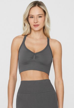 Mujer rubia con un sujetador deportivo gris oscuro y leggings de talle alto a juego, de pie frente a un fondo claro y liso.