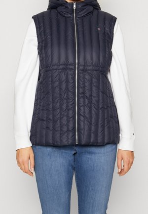 Kvinde iført en mørkeblå quiltet ærmeløs vest med frontlynlås over en hvid langærmet skjorte og blå jeans.