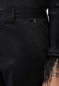 Melnas augsta vidukļa bikses ar sānu kabatām, valkātas kopā ar melnu adītu garroku topu, redzama arī tetovēta roka ar bārkstīm uz aprocēm.