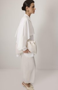 Hvid lagdelt bluse med gennemsigtige ærmer, parret med en matchende lang nederdel. Holder en blød beige clutch og har lyse højhælede sko på.