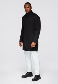 Zwarte wolmixjas met een hoge kraag en zijzakken met rits, gecombineerd met witte broek en zwarte veterschoenen. Gladde textuur.