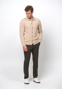 Beige Strickjacke mit Kragen und Fronttasche, mit dunklen Knöpfen. Kombiniert mit dunklen Hosen und weißen Sneakers. Glatte Textur.