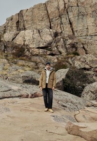 Jachetă puffer bej cu glugă, hanorac gri, pantaloni negri și încălțăminte deschisă purtate de o persoană care stă pe un teren stâncos.