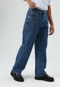 Jeans en denim bleu avec une coupe décontractée, des surpiqûres contrastées et cinq poches. Associés à des chaussures à plateforme noires avec des lacets.