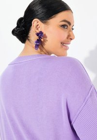 Maglione lilla con un design a maglia testurizzato, caratterizzato da un ampio scollo e una vestibilità ampia. Orecchini con strati di petali viola che aggiungono interesse visivo.