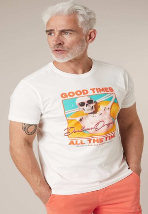 Homme aux cheveux blancs et à la barbe, portant un t-shirt blanc avec un graphique coloré de squelette et un short orange, regardant sur le côté.