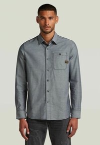Chemise en chambray bleu à manches longues, devant boutonné, col, poche poitrine unique et étiquette sur la poche. Portée avec un jean noir déchiré.