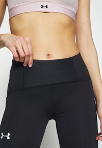 Femme portant un soutien-gorge de sport rose clair et un legging noir taille haute Under Armour, ajustant la taille avec la main gauche.
