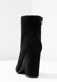 Bottine noire en daim avec un talon bloc haut, dotée d'un bout rond et d'une fermeture éclair latérale pour un enfilage facile. Texture lisse avec une finition mate.