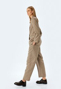 Traje beige a medida con un ajuste holgado, que incluye una blazer y pantalones de pierna ancha, complementado con zapatos negros brillantes de cordones.
