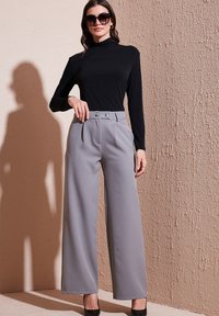 Black long-sleeve top paired with high-waisted, wide-legged gray trousers featuring button accents; model wears black heels and sunglasses.
