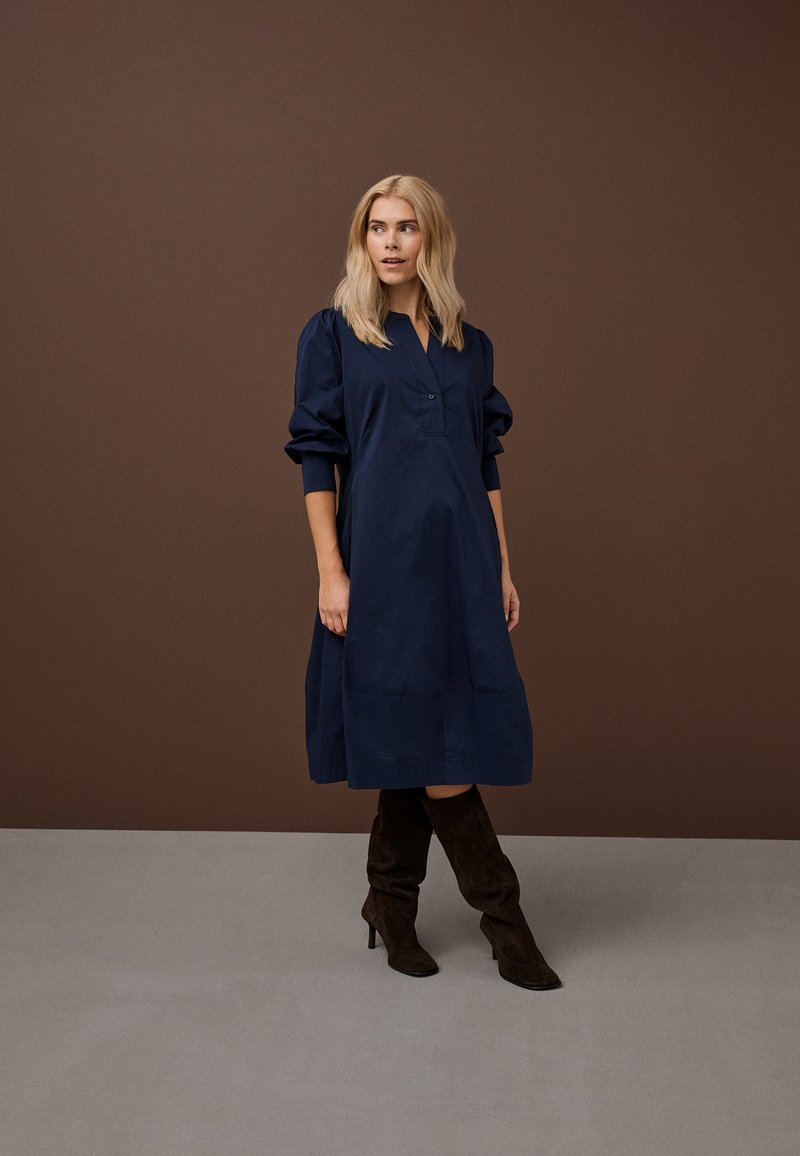 Plavolaska ženska v ohlapni temno modri midi obleki z napihnjenimi rokavi in temno rjavi škornji z višino do kolen ter peto stoji na sivi površini.