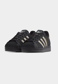 Μαύρα sneakers Adidas Superstar με εκρού ρίγες και σχεδιασμό με μύτη σε σχήμα κοχύλι, τοποθετημένα δίπλα δίπλα σε λευκό φόντο.