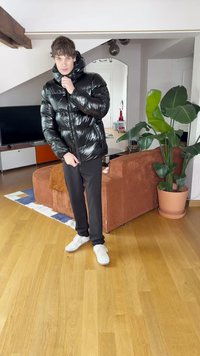 Casaco preto brilhante acolchoado com gola alta, com fecho éclair. Combinado com calças pretas e ténis brancos, de pé numa sala com plantas.