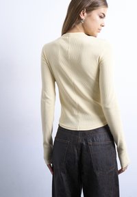 Γυναίκα με μακριά μαλλιά, που φοράει μπεζ ριγωτό μακρυμάνικο τοπ και σκούρο υφασμάτινο παντελόνι, εμφανιζόμενη από πίσω πάνω σε απλή φόντο.