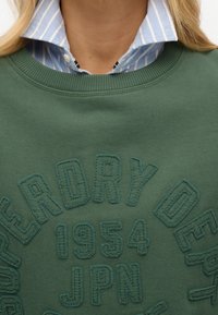 Sudadera verde con texto bordado "SUPERDRY DEPT" y "1954 JPN". Superpuesta sobre una camisa de cuello a rayas azules y blancas.