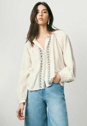 Woman wearing a loose cream blouse with black embroidery and wide-leg light blue jeans, standing with one hand in pocket against plain background.