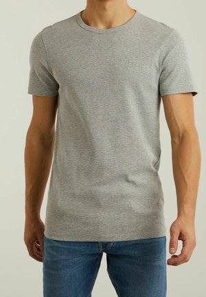 Mężczyzna w prostym jasnoszarym t-shircie z krótkim rękawem i niebieskich dżinsach, stoi z rozluźnionymi ramionami opuszczonymi wzdłuż ciała na neutralnym tle.