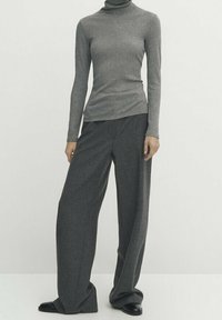 Personne portant un col roulé gris ajusté et un pantalon large gris foncé ample, debout devant un fond clair uni.