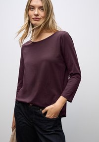 Paarse langemouwen shirt met een bootnek, zachte textuur en een licht ontspannen pasvorm, gecombineerd met donkere spijkerbroek.