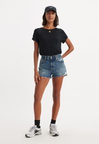 Camiseta negra, pantalones cortos de mezclilla desgastados de cintura alta y zapatillas grises. Gorra negra con el logo de "Levi's". Atuendo casual de verano.