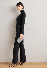 Schwarzes Rollkragenoberteil, hochgeschnittene schwarze Jeans und schwarze, verzurrte High Heels; hält eine kleine Tasche mit silbernen Akzenten; Seitenansicht.